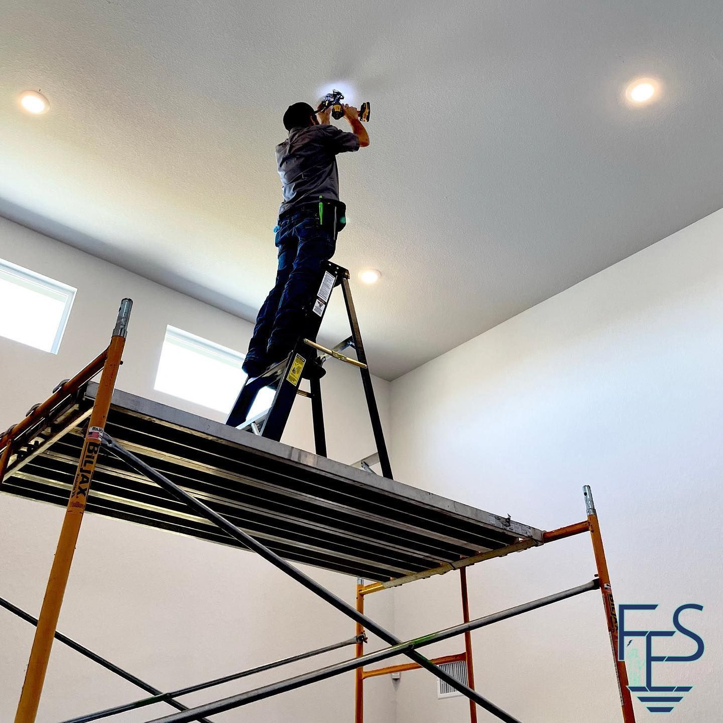 A person installs a light fixture on a ceiling, standing on a scaffold in a room with white walls.